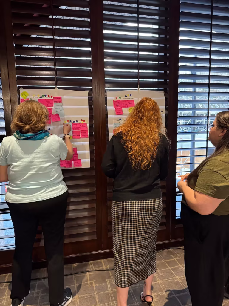 Three people standing in front of large wooden window shutters, placing colorful sticky notes on posters during a collaborative workshop activity