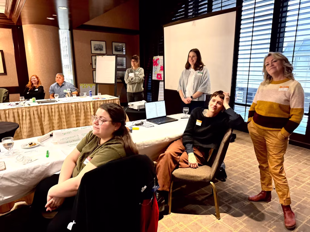 A group of professionals in a meeting room with tables, a projector screen, and a whiteboard