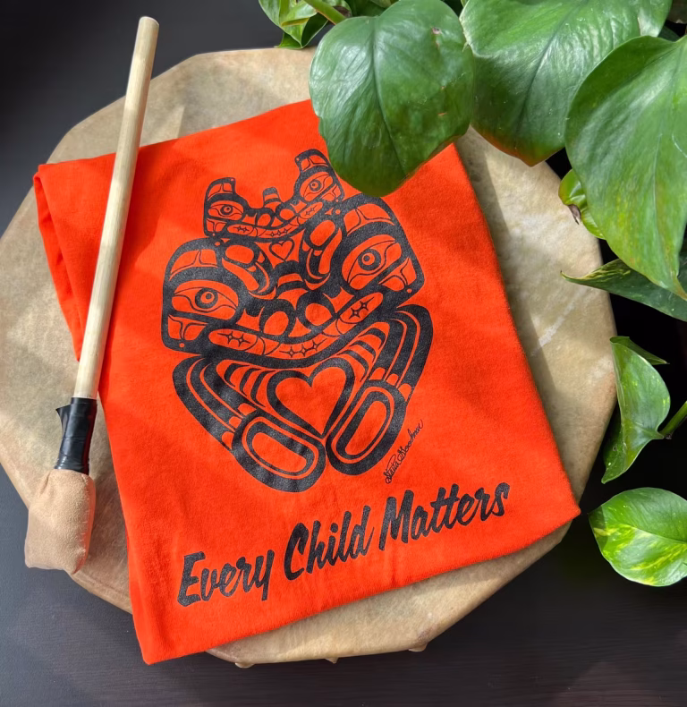 An orange t-shirt with the text Every Child Matters and a black formline mother bear design lies on a drum next to a drumstick
