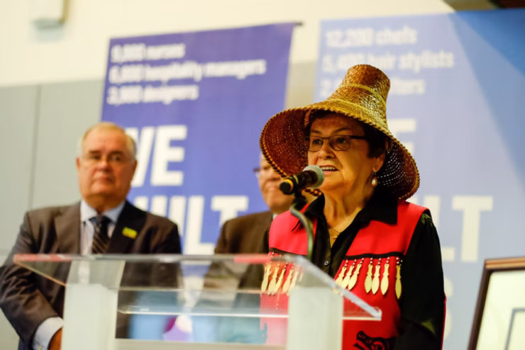 Elder Deanna George wearing a traditional woven cedar hat and a red and black regalia vest speaking into a microphone at a podium