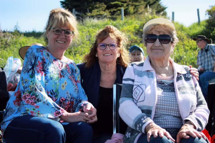 Three women sitting close together and smiling for a photo during a sunny outdoor community gathering