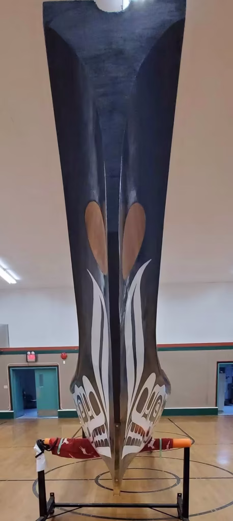 A tall, carved K’ómoks First Nation canoe standing upright in a gymnasium