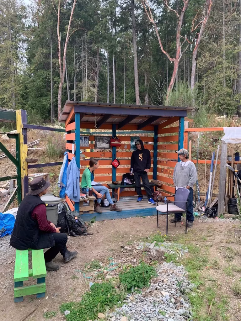 A small group of people gathered around a bright orange and blue wooden shelter in a forested area