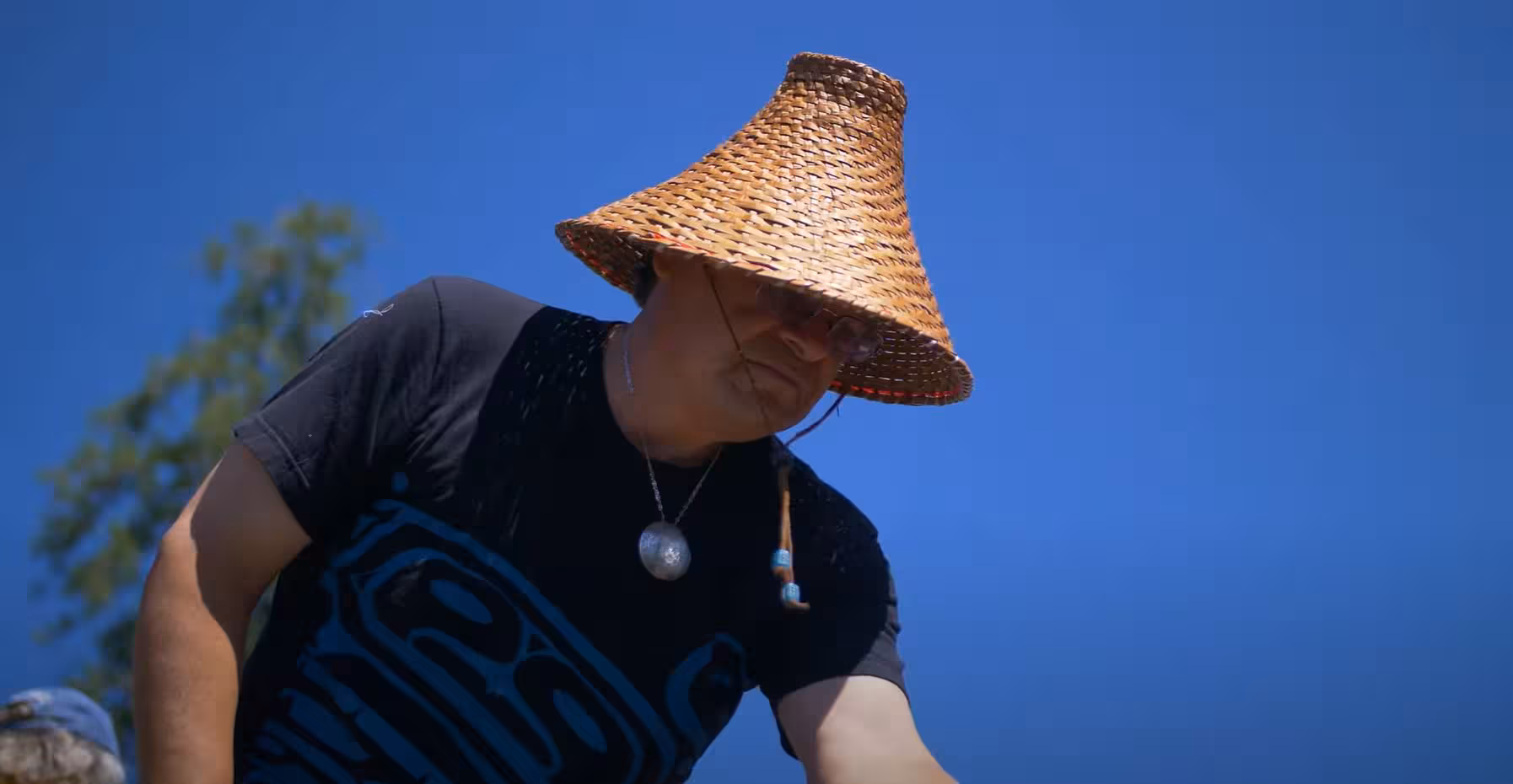 A T'Souke man wearing a traditional woven cedar hat and a black shirt with a blue Indigenous design looks downward against a clear blue sky