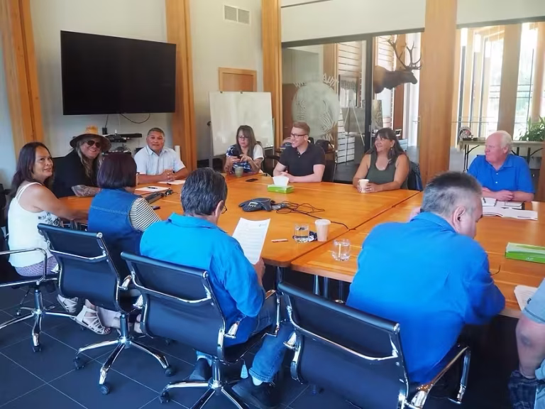 A diverse group of people are seated around a large wooden conference table in a modern meeting room.
