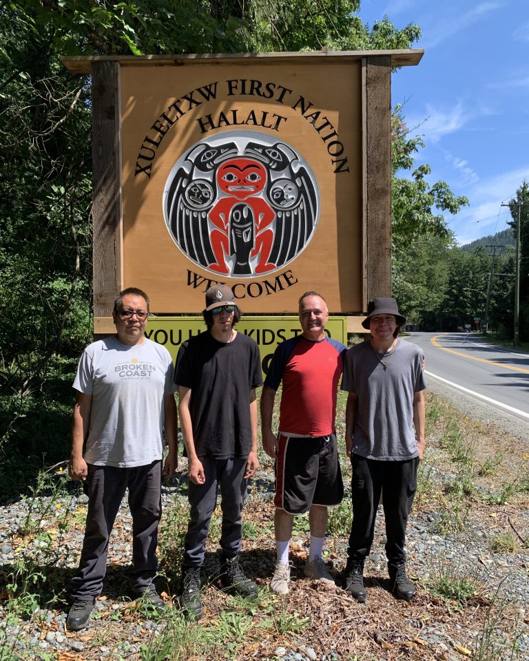 Four men stand in front of a large wooden sign for XULEELTXW FIRST NATION HALALT.