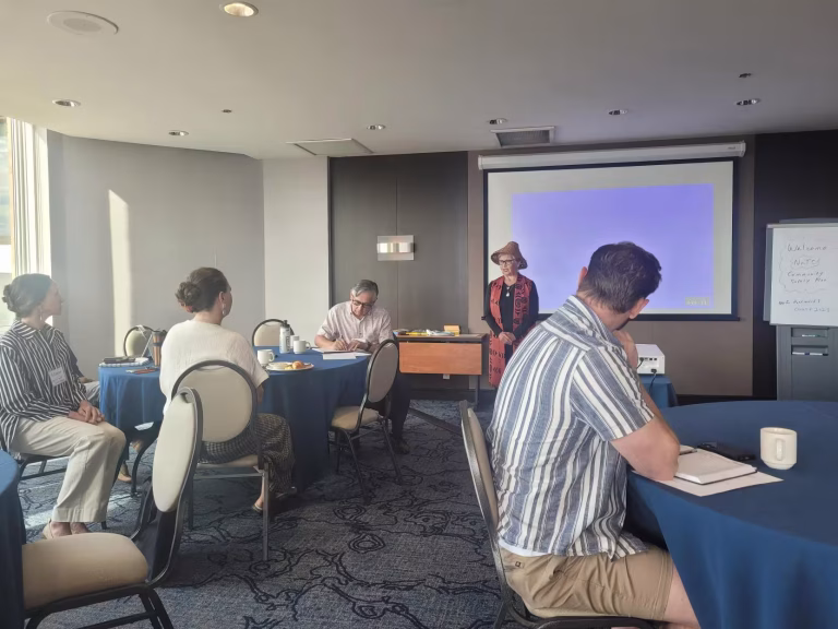 A woman in a hat presents to a group of people seated at round tables in a conference room.