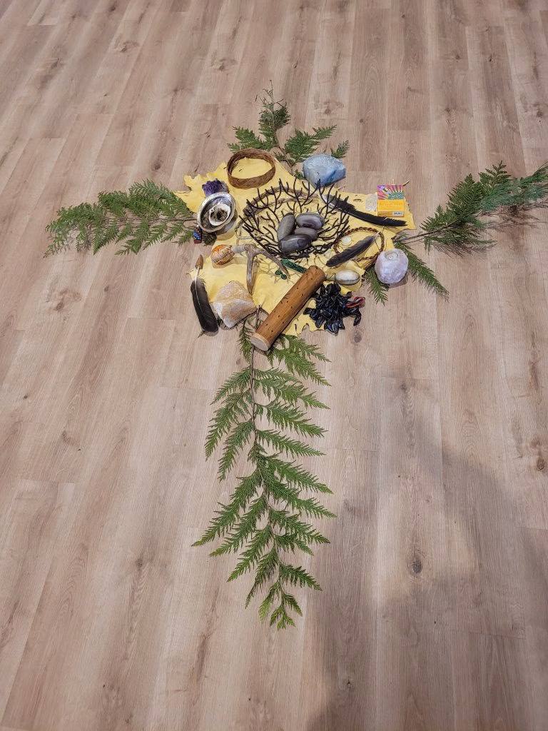 A cross shape is made with natural items including feathers, stones, crystals, and greenery on a wooden floor.