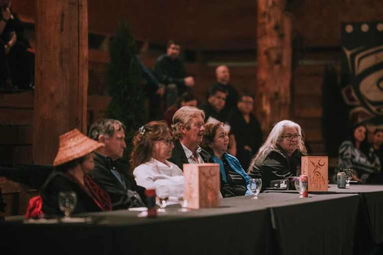 A diverse group of people sit at a table in a dimly lit room with wooden beams, listening attentively.