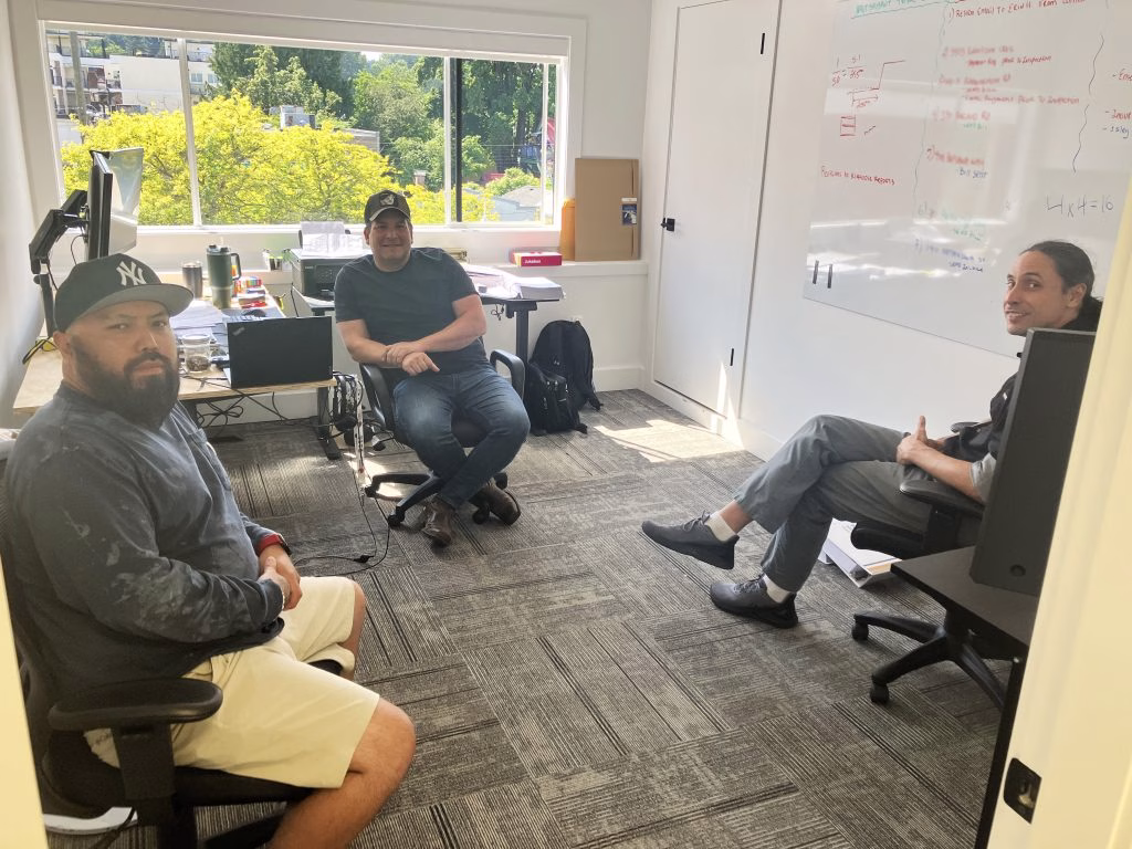 Three men sit in chairs in a bright office with a whiteboard and a large window.