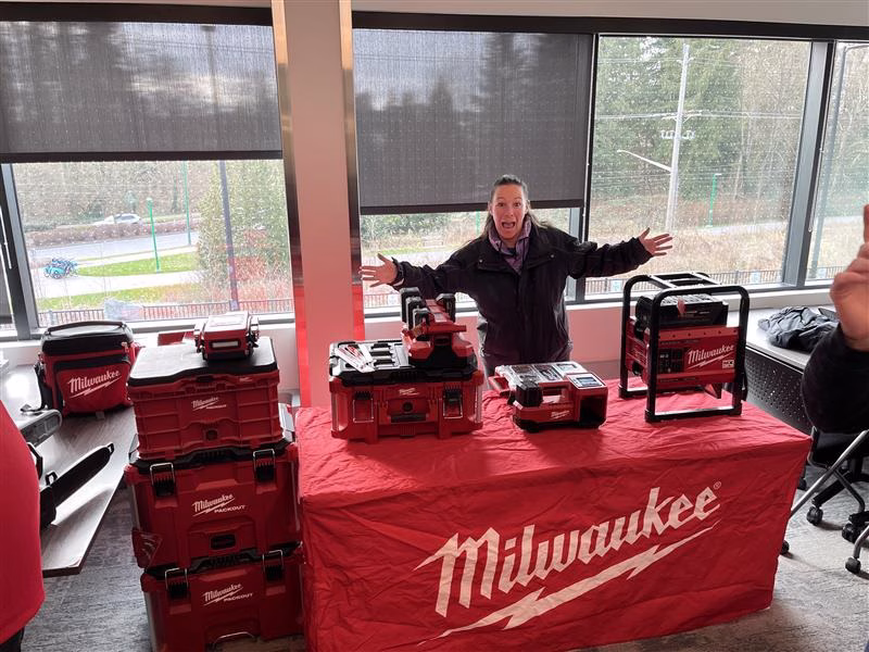 Woman with arms outstretched stands behind a table displaying red Milwaukee toolboxes and equipment.