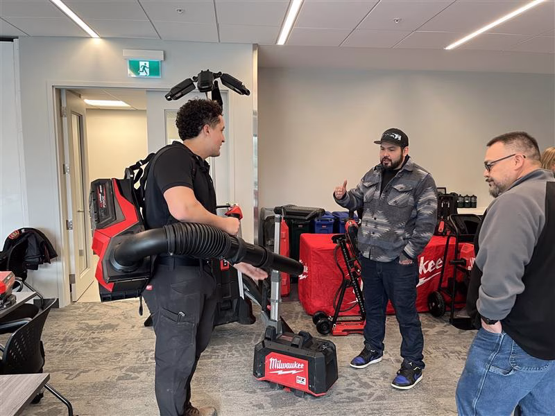 Men in a room with Milwaukee tools, one wearing a backpack leaf blower.