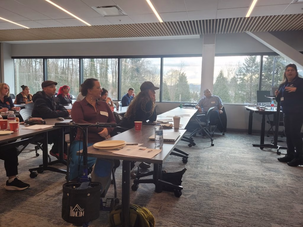 A group of people sit at tables in a modern conference room, listening to a presenter.