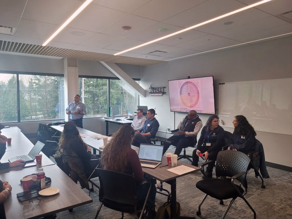 A man stands and presents to a group of people seated around tables in a modern conference room.