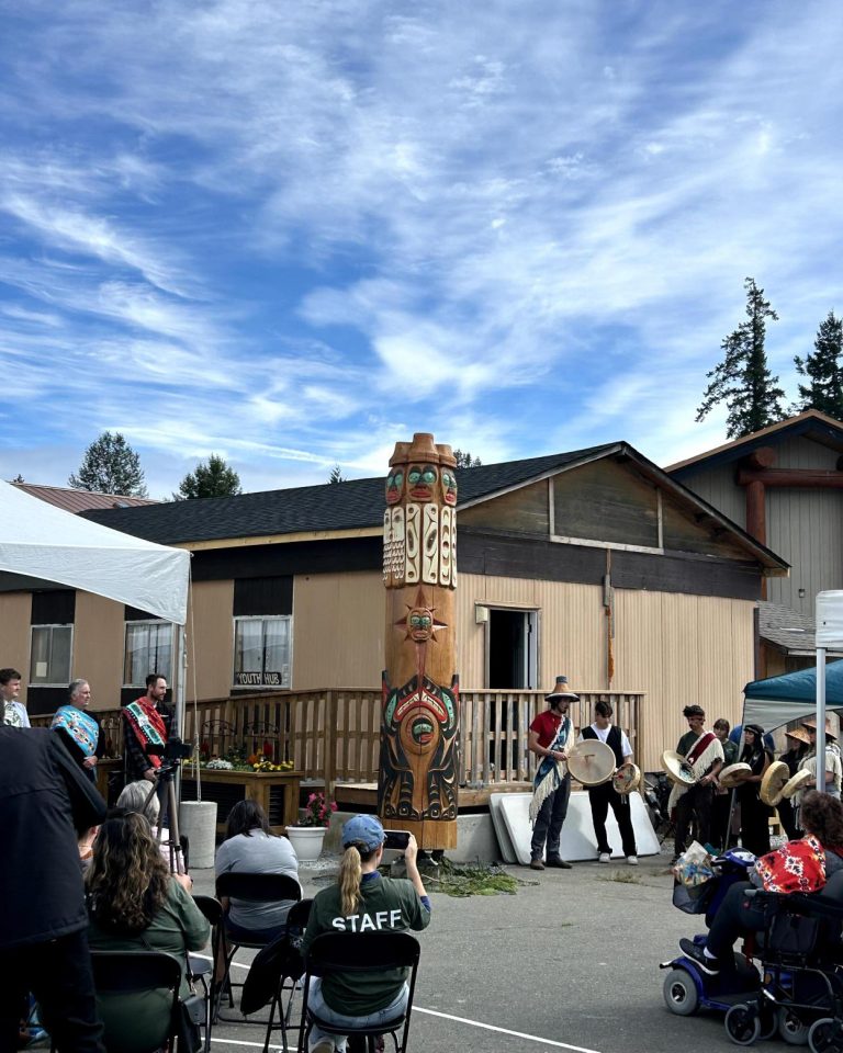 People in traditional regalia drum and sing by a totem pole in front of a building.