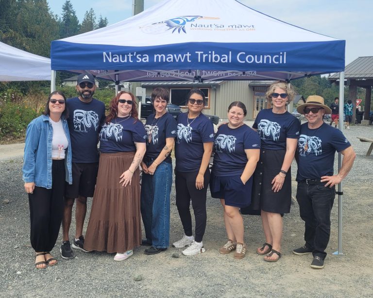 A group of eight people stand in front of a blue tent with "Naut'sa mawt Tribal Council" written on it.