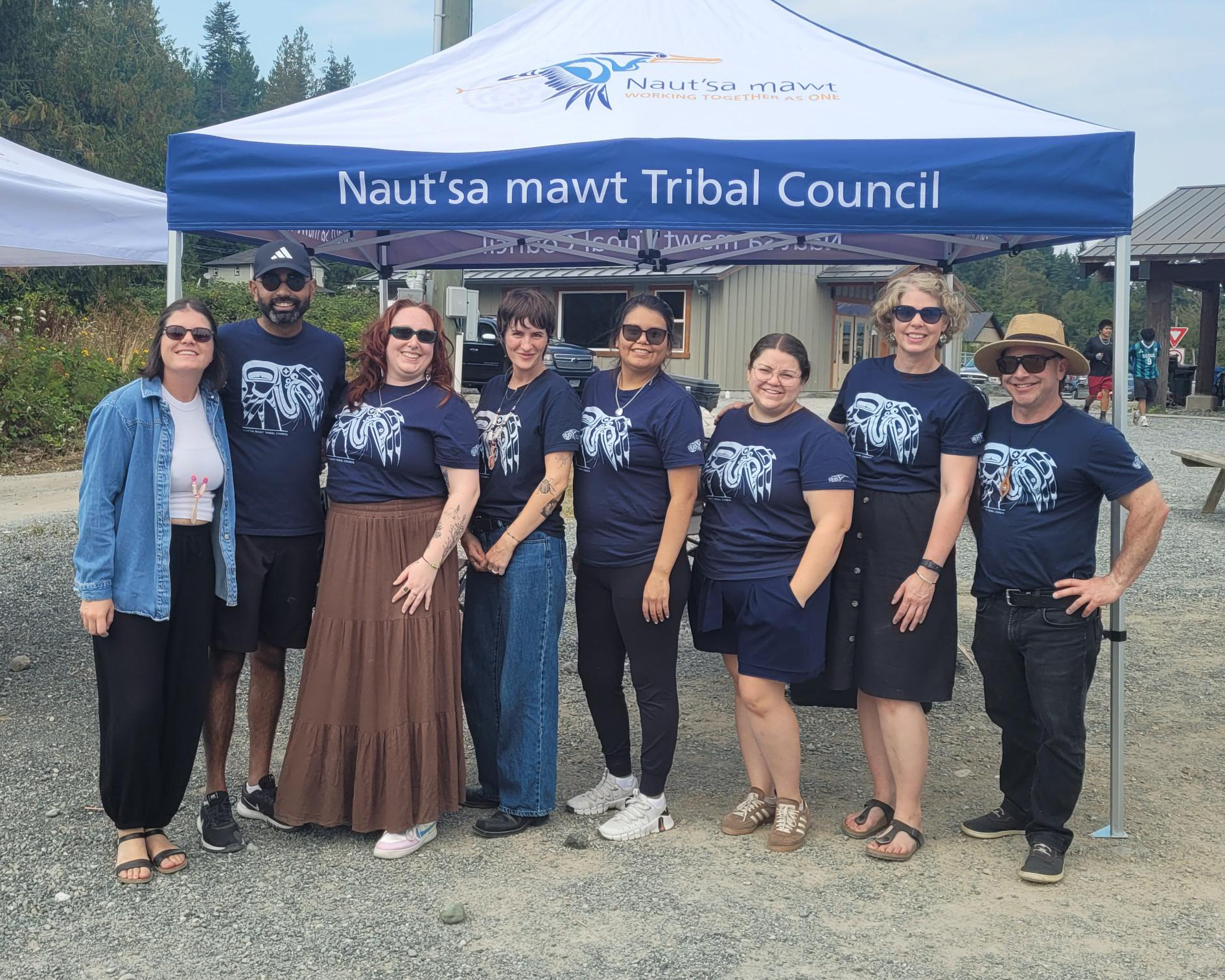 A group of eight people stand in front of a blue tent with "Naut'sa mawt Tribal Council" written on it.