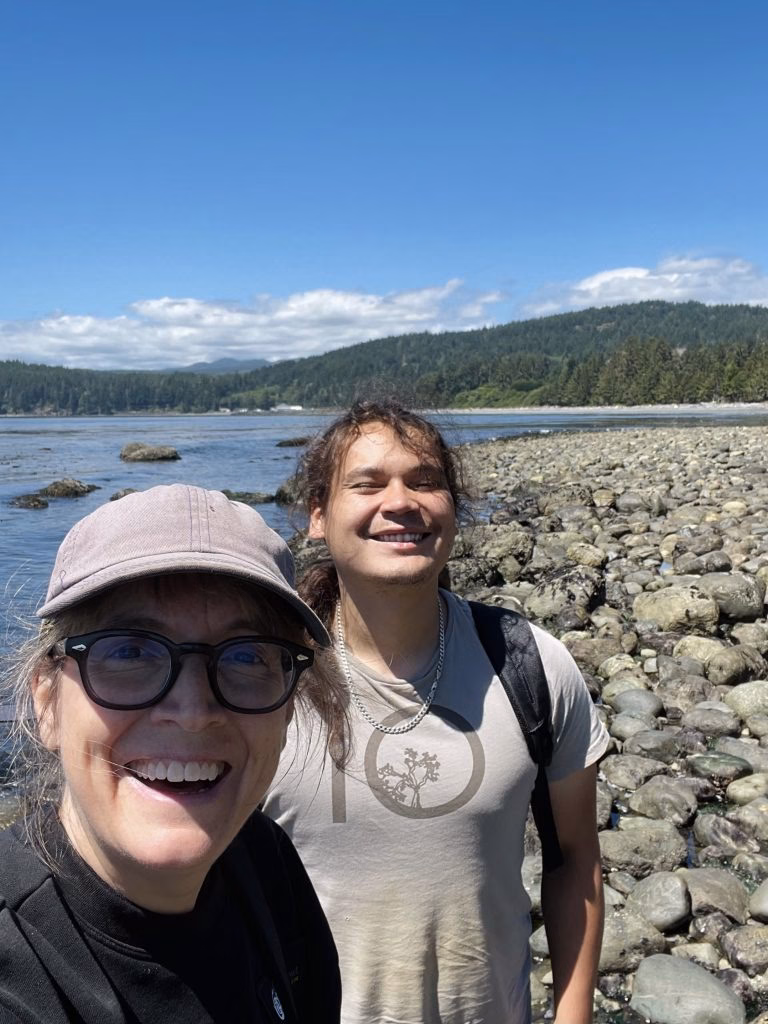 Two people smile for a selfie on a rocky beach with a forested coastline and cloudy sky in the background.