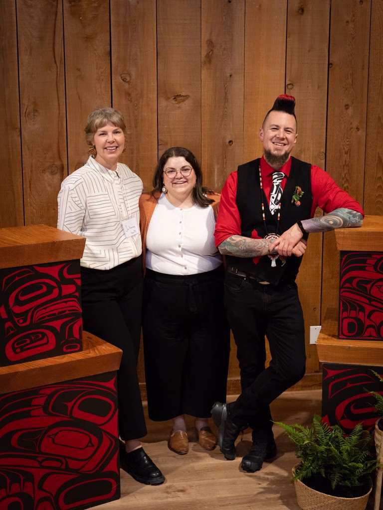 Three people pose in front of a wooden wall, with Indigenous art boxes on either side.