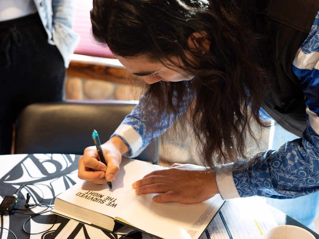 Person writing in a book with text "WE SURVIVED THE NIGHT" on a table with a black and white patterned cloth.