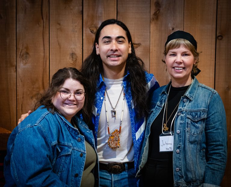 Three smiling people in denim jackets pose against a wood-paneled background.