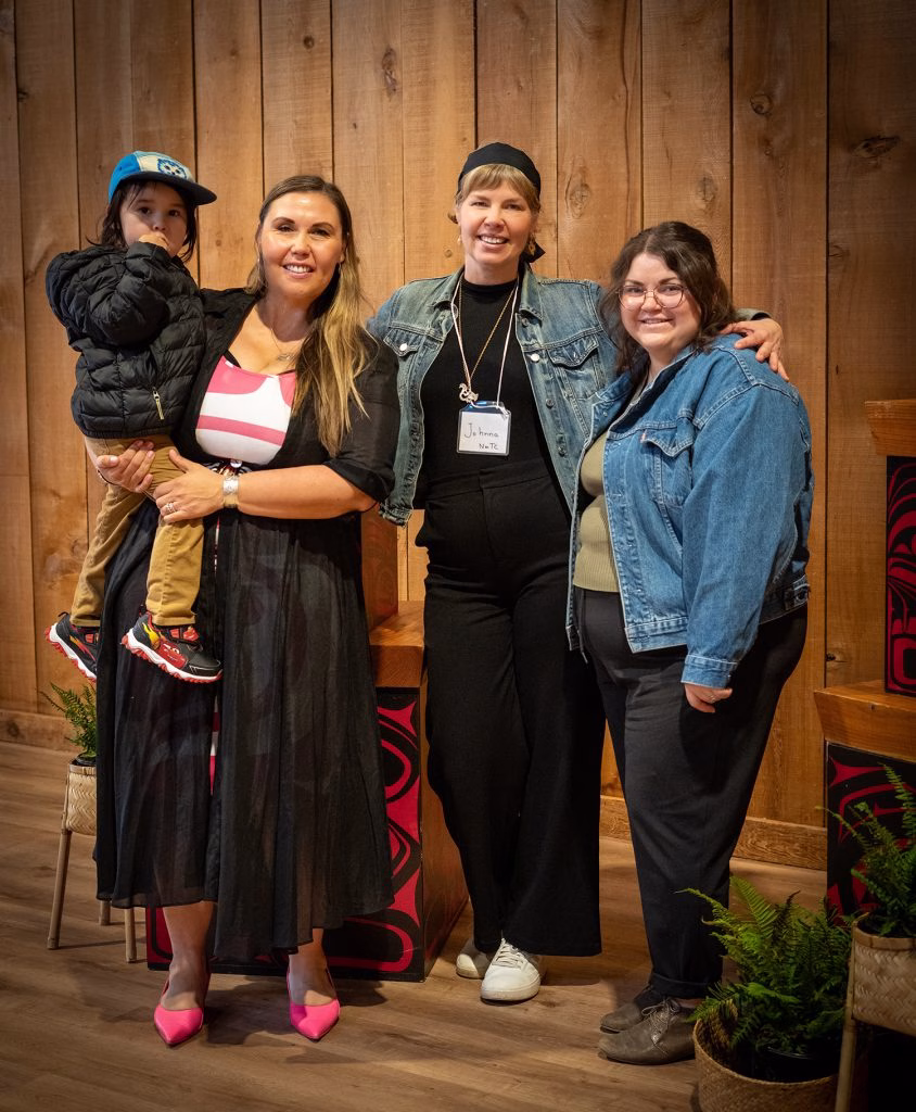 A woman holds a young boy while standing with two other women in front of a wooden wall.
