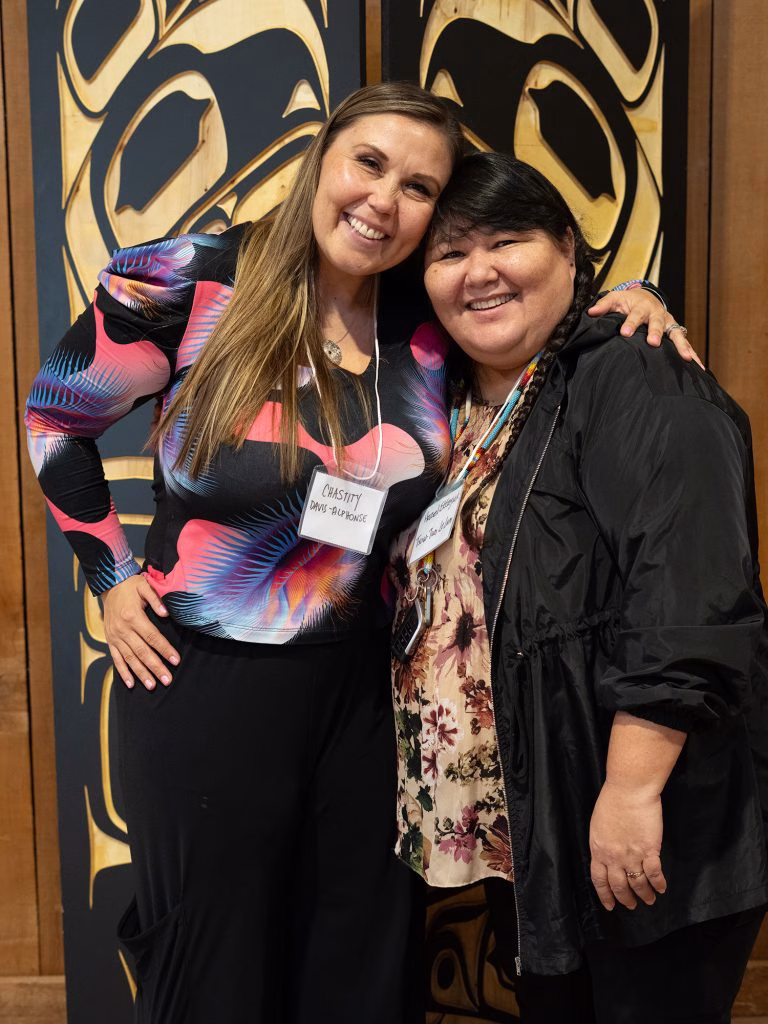 Chastity Davis-Alphonse and Hazel Littlelight pose together in front of a carved wooden panel.