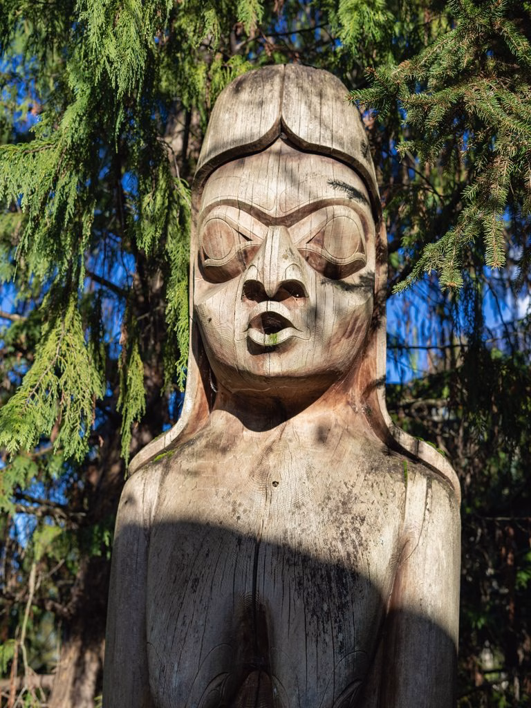 Close-up of a weathered wooden totem pole with a carved face, set against green trees and blue sky.