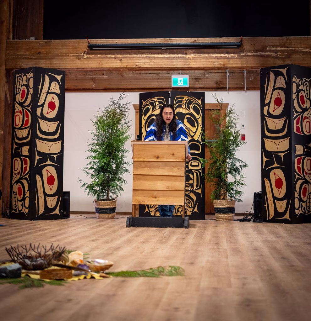 A person stands at a wooden podium in front of a large, intricately designed backdrop, flanked by two tall, decorated columns and potted plants.