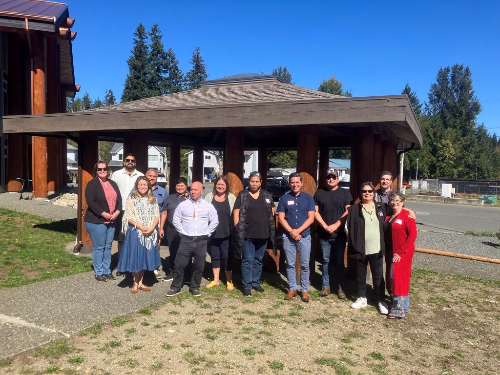 A diverse group of people stands in front of a wooden building with a covered walkway on a sunny day.