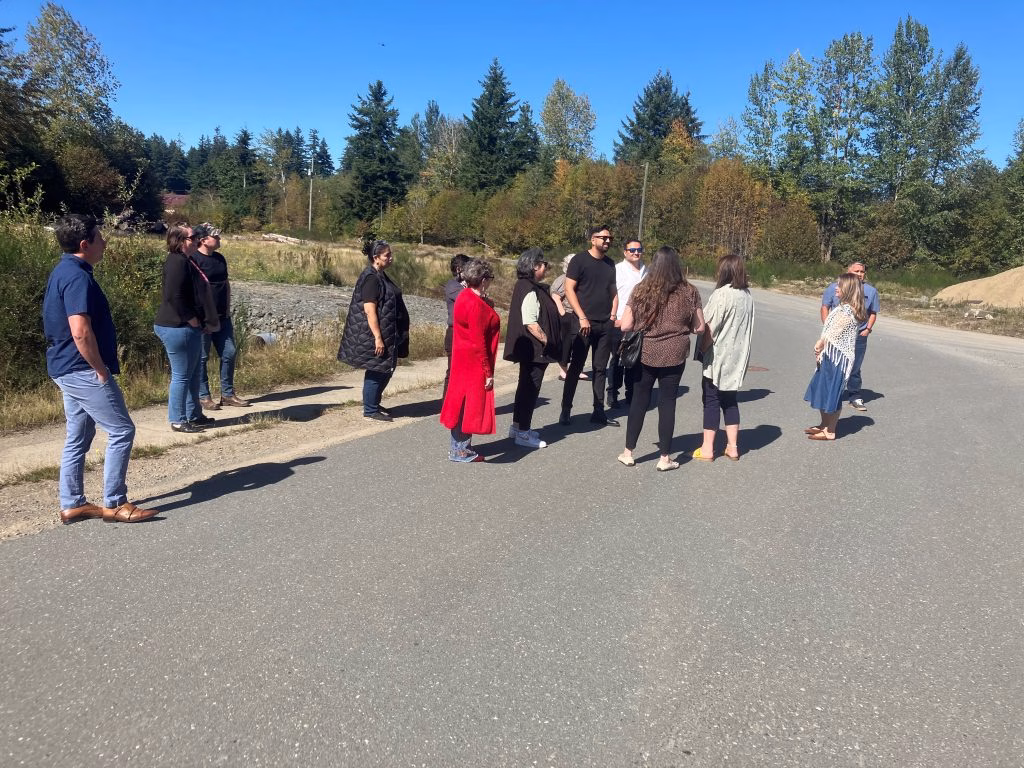 A diverse group of people stand gathered on a paved road, surrounded by trees and a clear blue sky.