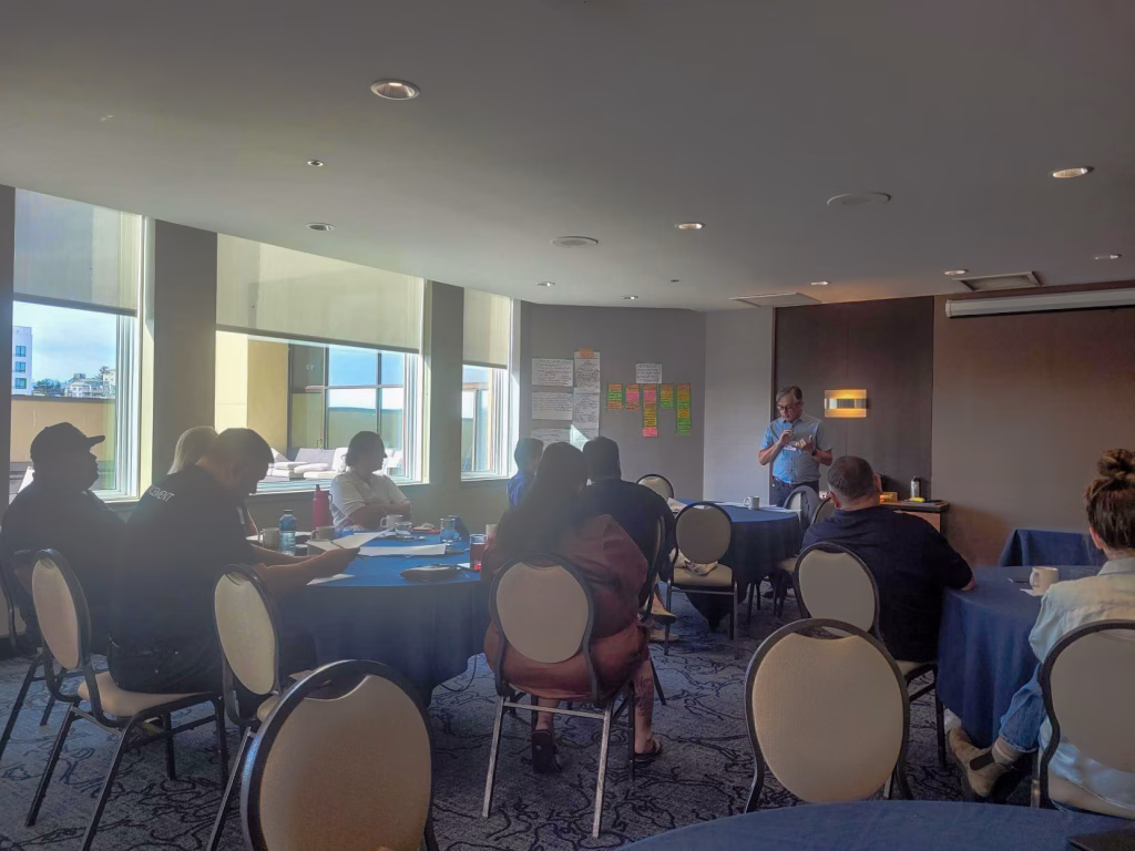 People sit at tables in a conference room while a man stands and presents to them.